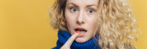 Isolated shot of amazed beautiful female with curly blonde hair, keeps fore finger on lower lip, listens someone attentively, dressed in blue sweater, isolated over yellow background. Emotions