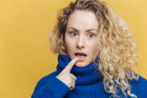 Isolated shot of amazed beautiful female with curly blonde hair, keeps fore finger on lower lip, listens someone attentively, dressed in blue sweater, isolated over yellow background. Emotions